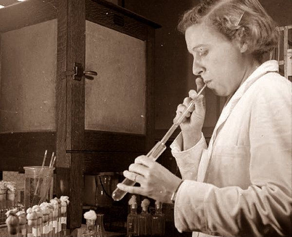 A black and white image of a woman in the lab using her mouth to pipette cells from one test tube to another.